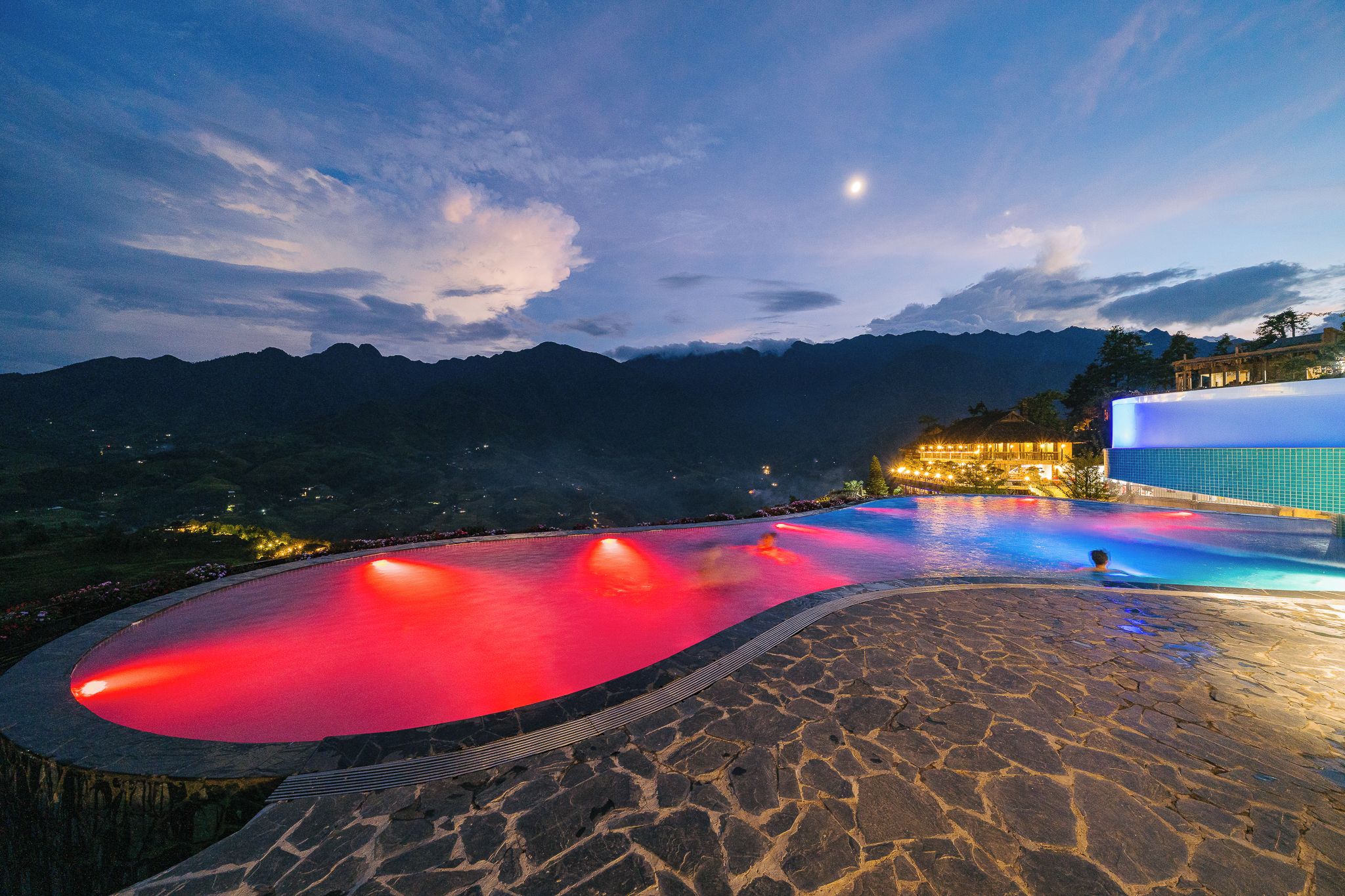 A swimming pool with a stone deck, surrounded by mountains and a lit building.