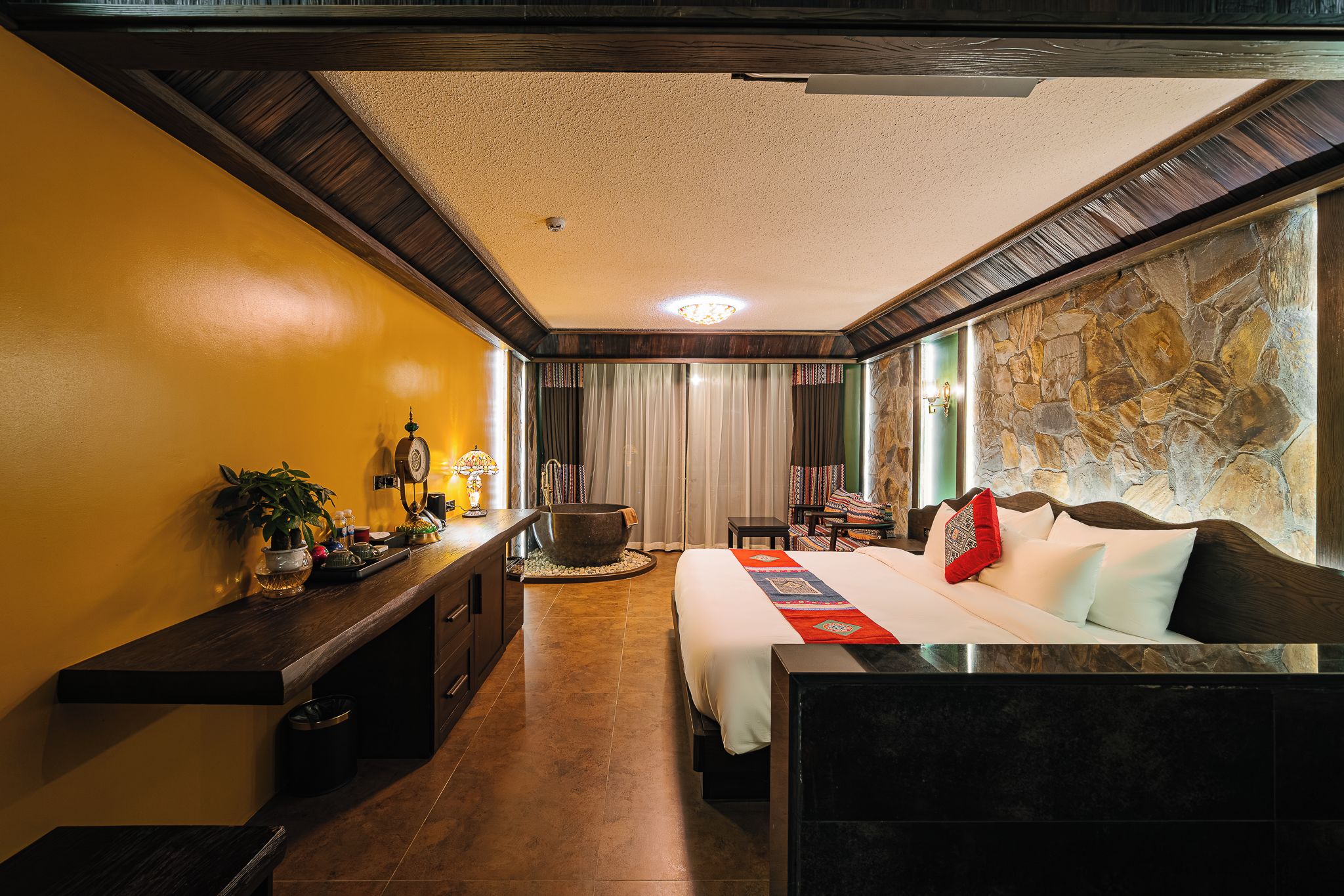 Habitació d'hotel amb llit, taulella de nit, làmpada, cortines i una paret de pedra.
