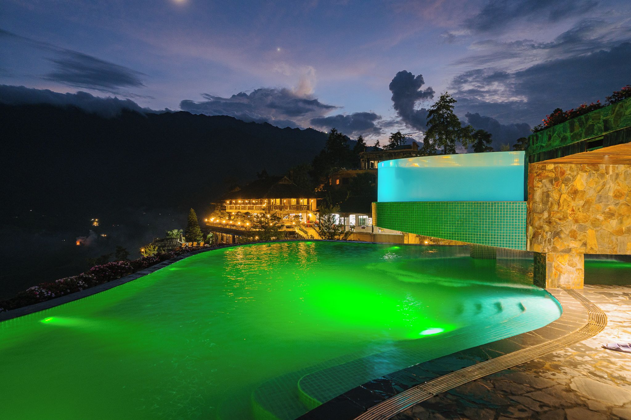 Una piscina nocturna de tono verdoso, rodeada por un muro de piedra y un edificio con luces.