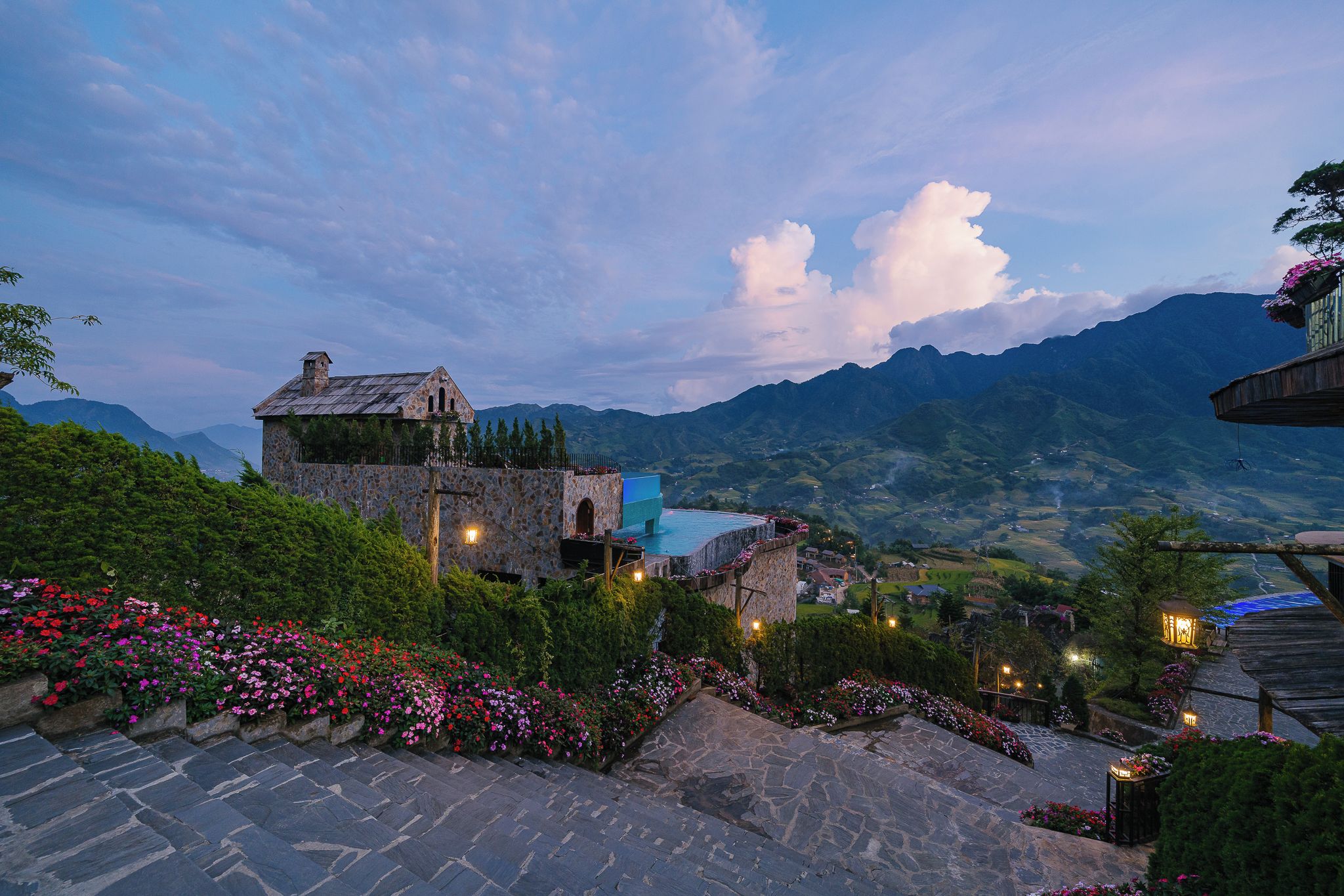 A stone building with a pool, surrounded by a garden with flowers and a view of mountains.