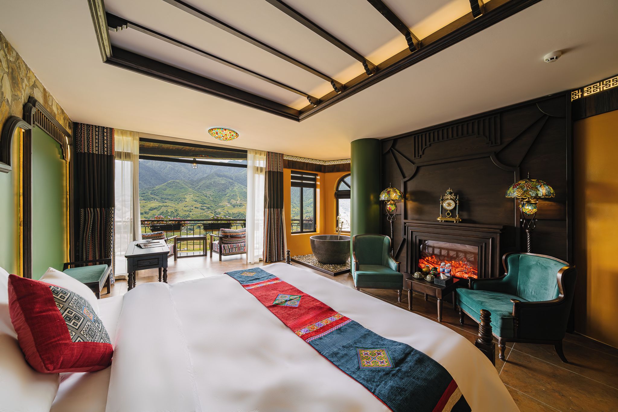 A hotel room with a large bed, a fireplace, a balcony with a view of mountains, and a dining area.