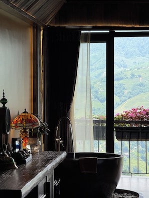 Una habitación con un ventanal grande, un escritorio de madera, una lámpara y vistas a colinas verdes.