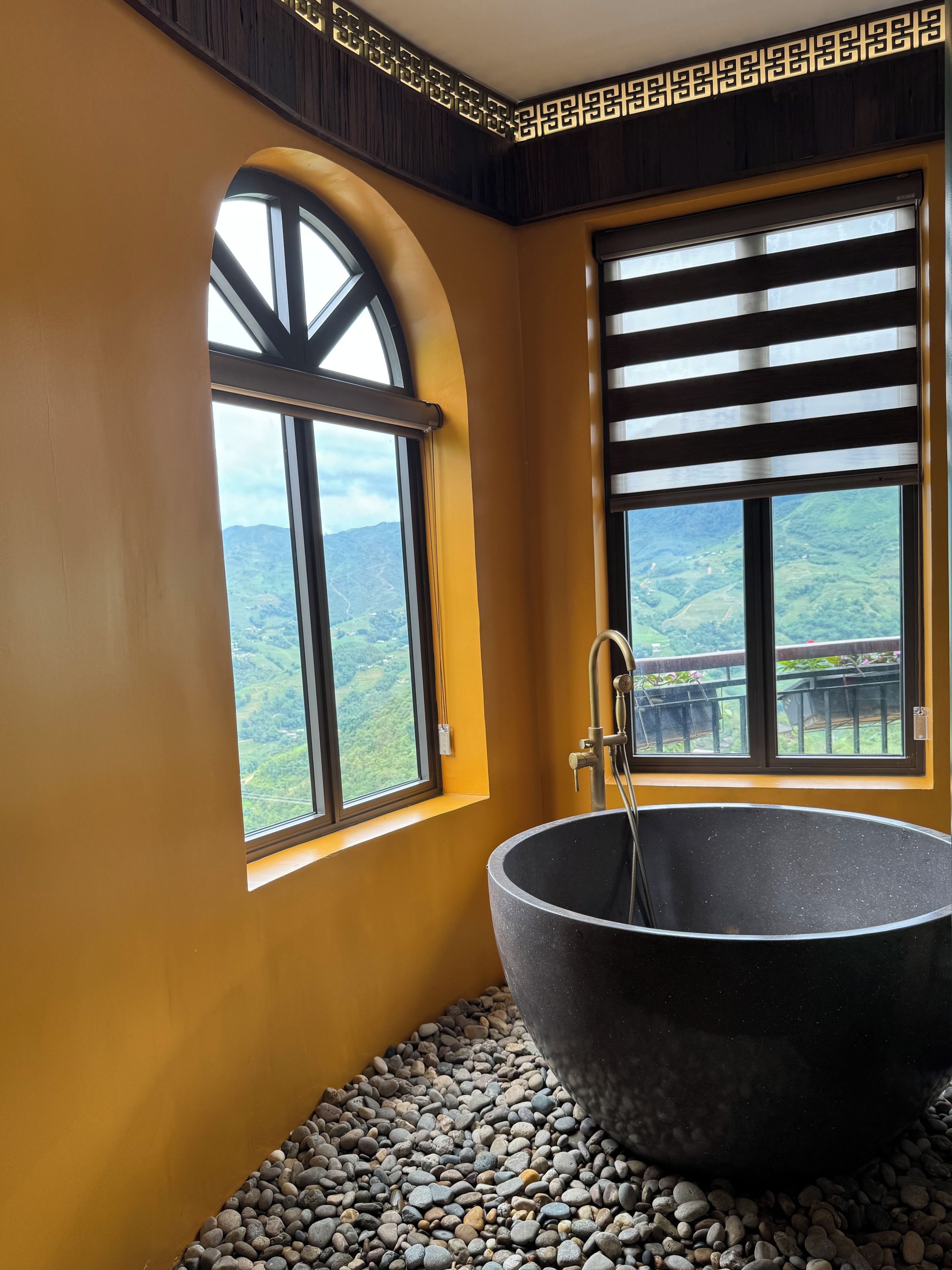 Un baño con una bañera negra grande, dos ventanas con vistas a colinas verdes y un techo decorativo de madera.