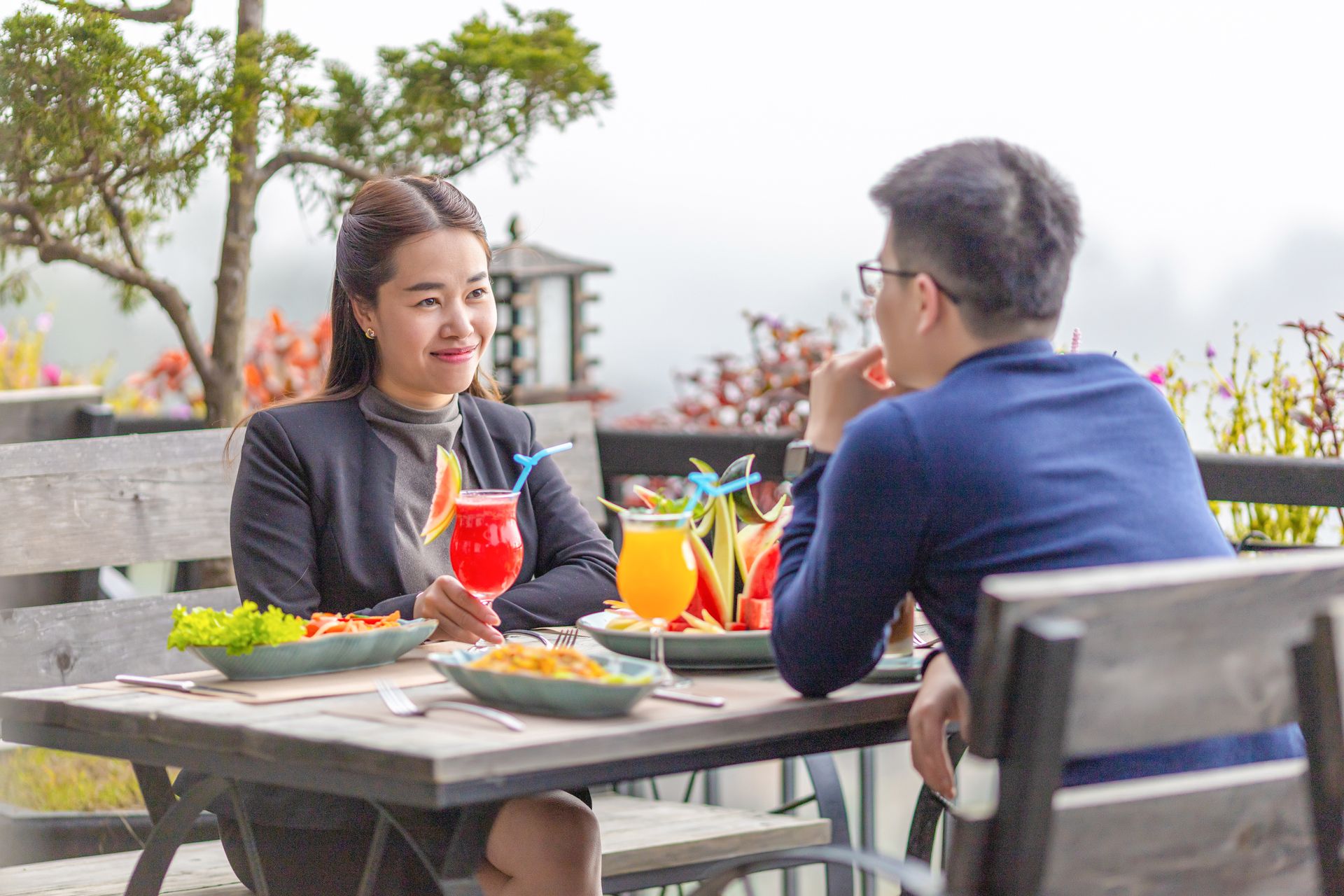 Dos personas sentadas en una mesa al aire libre, con platos de comida y bebidas.