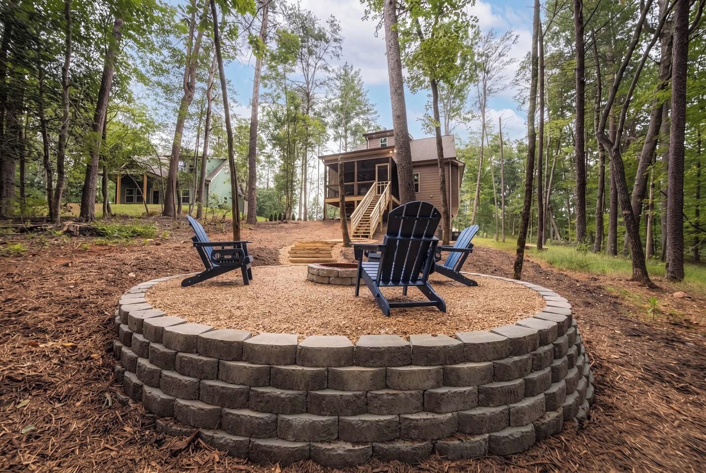 A circular seating area with Adirondack chairs, a fire pit, and a wooden deck leading to a cabin in a forested setting.