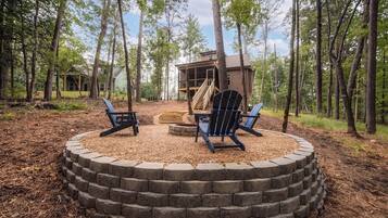 A circular seating area with Adirondack chairs, a fire pit, and a wooden deck leading to a cabin in a forested setting.