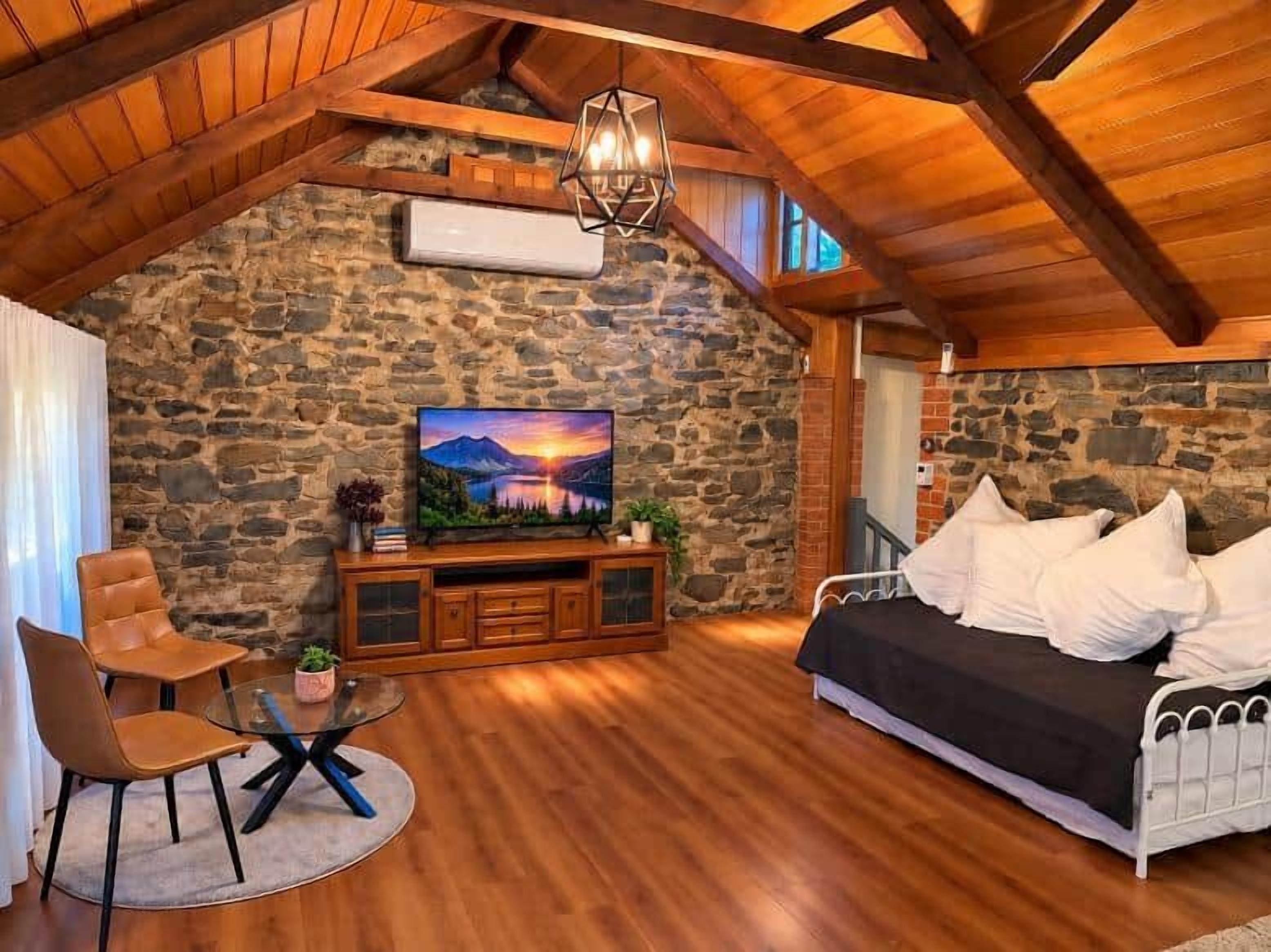 A cozy room with a stone wall, wooden ceiling beams, a bed, a TV, and a seating area with a table and chairs.