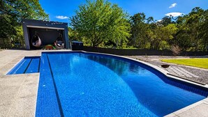 A swimming pool with a clear blue water surface, surrounded by a paved deck and a grassy area. - Blumberg Mews - Stunning Adelaide Hills Retreat Near Barossa (Birdwood)