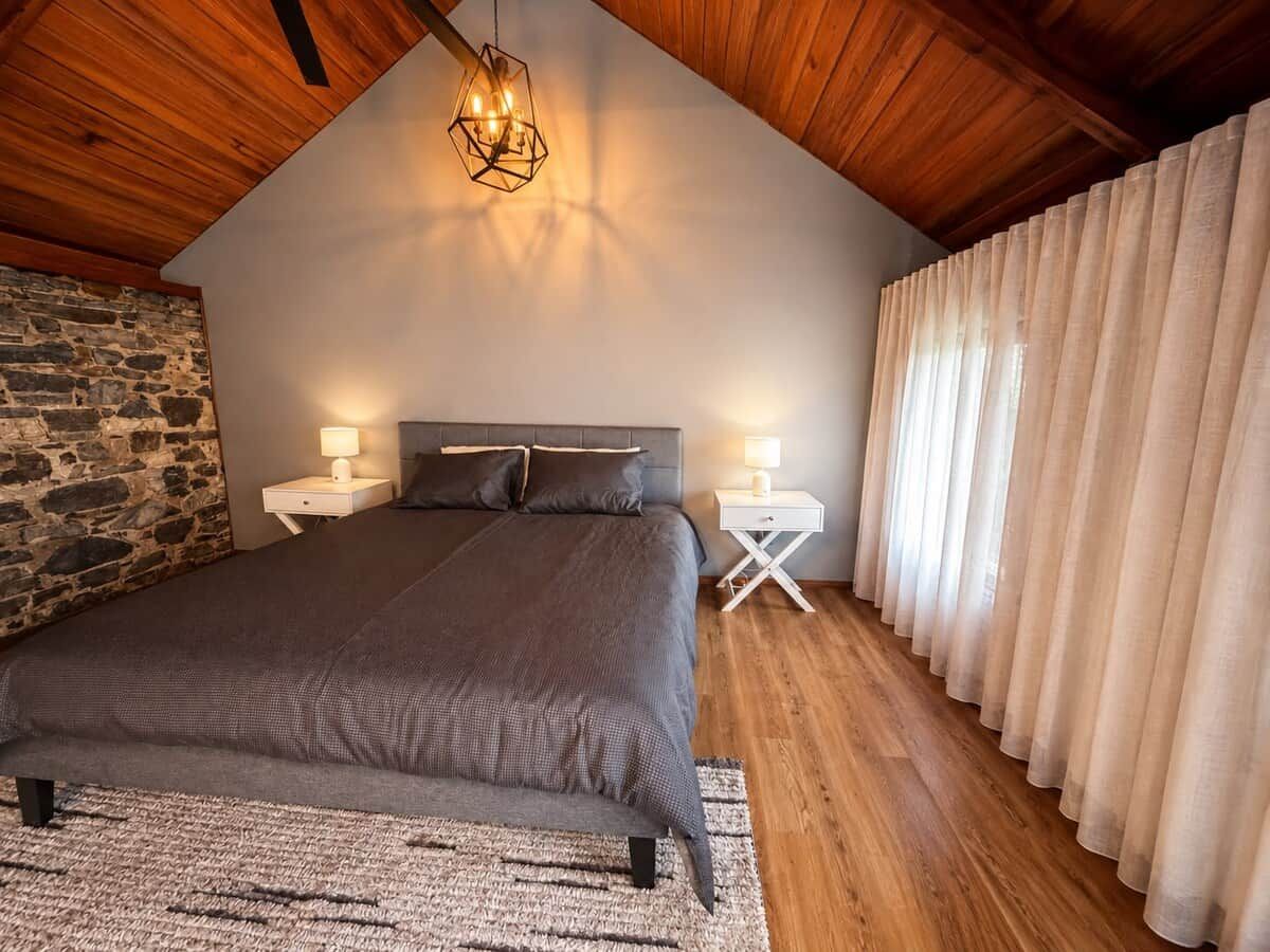 A bedroom with a sloped ceiling, a bed with a headboard, two bedside tables with lamps, a stone wall, and a window with curtains.