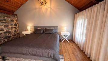 A bedroom with a sloped ceiling, a bed with a headboard, two bedside tables with lamps, a stone wall, and a window with curtains.