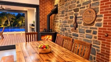 A wooden dining table with chairs, a bowl of fruit, and a stone wall with a fireplace.
