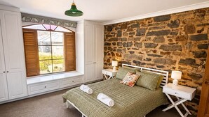A bedroom with a stone wall, a window with shutters, a bed with a green bedspread, and a nightstand with a lamp. - Blumberg Mews - Stunning Adelaide Hills Retreat Near Barossa (Birdwood)