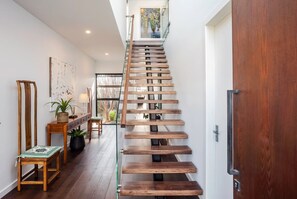 A modern staircase with glass railings and wooden steps leading to a second-floor landing. - The Clovelly House | Luxury Coastal Retreat (Holdfast Bay)