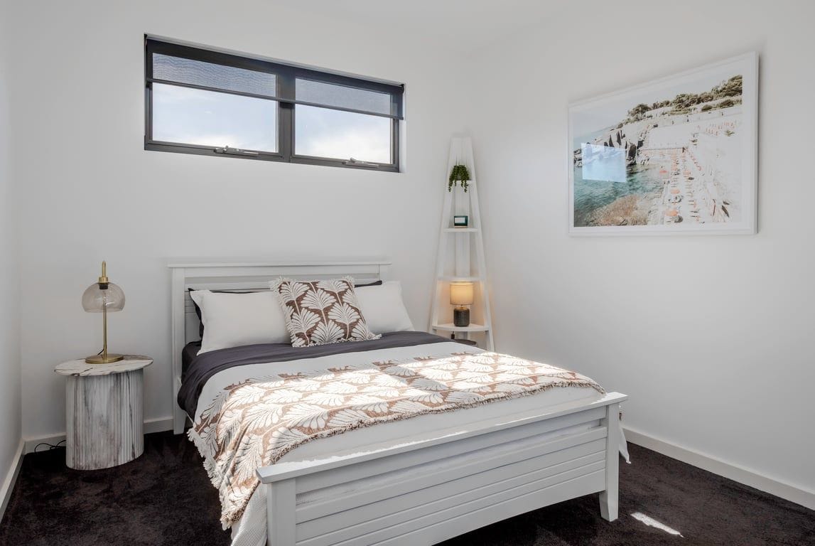 Quarto com cama, mesa de cabeceira, luminária, janela e prateleira na parede com uma planta.