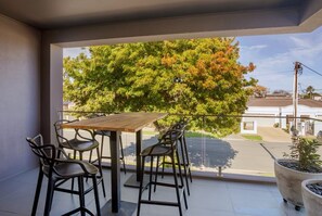 A balcony with a wooden table and chairs, overlooking a tree and a residential area. - The Clovelly House | Luxury Coastal Retreat (Holdfast Bay)