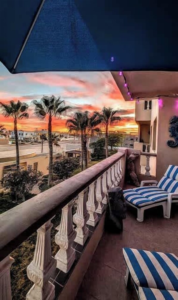 Un balcon offrant une vue sur un coucher de soleil, des palmiers et des chaises longues à rayures.