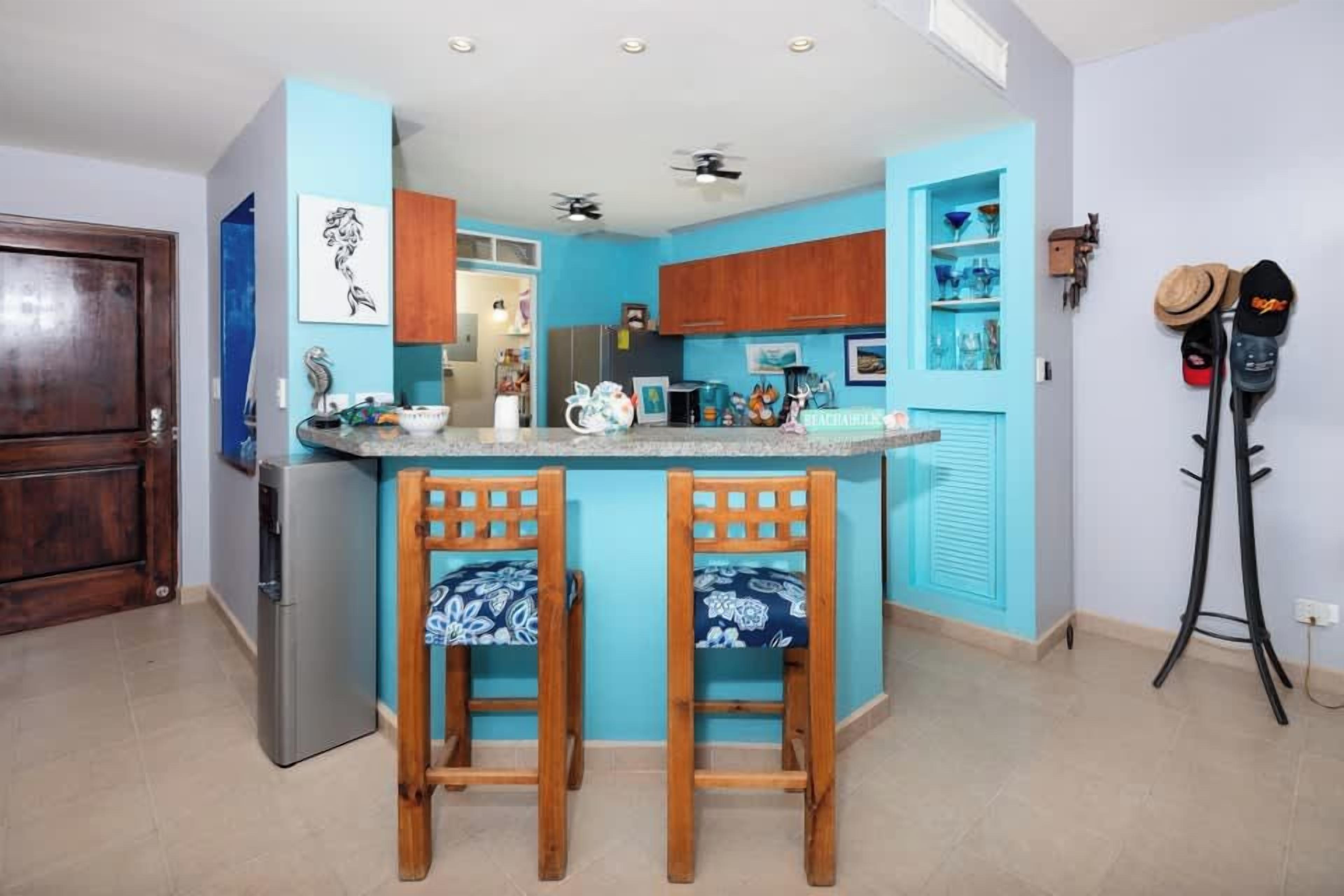 Cozinha com ilha central, cadeiras de madeira, geladeira e porta-chapéus fixado na parede.