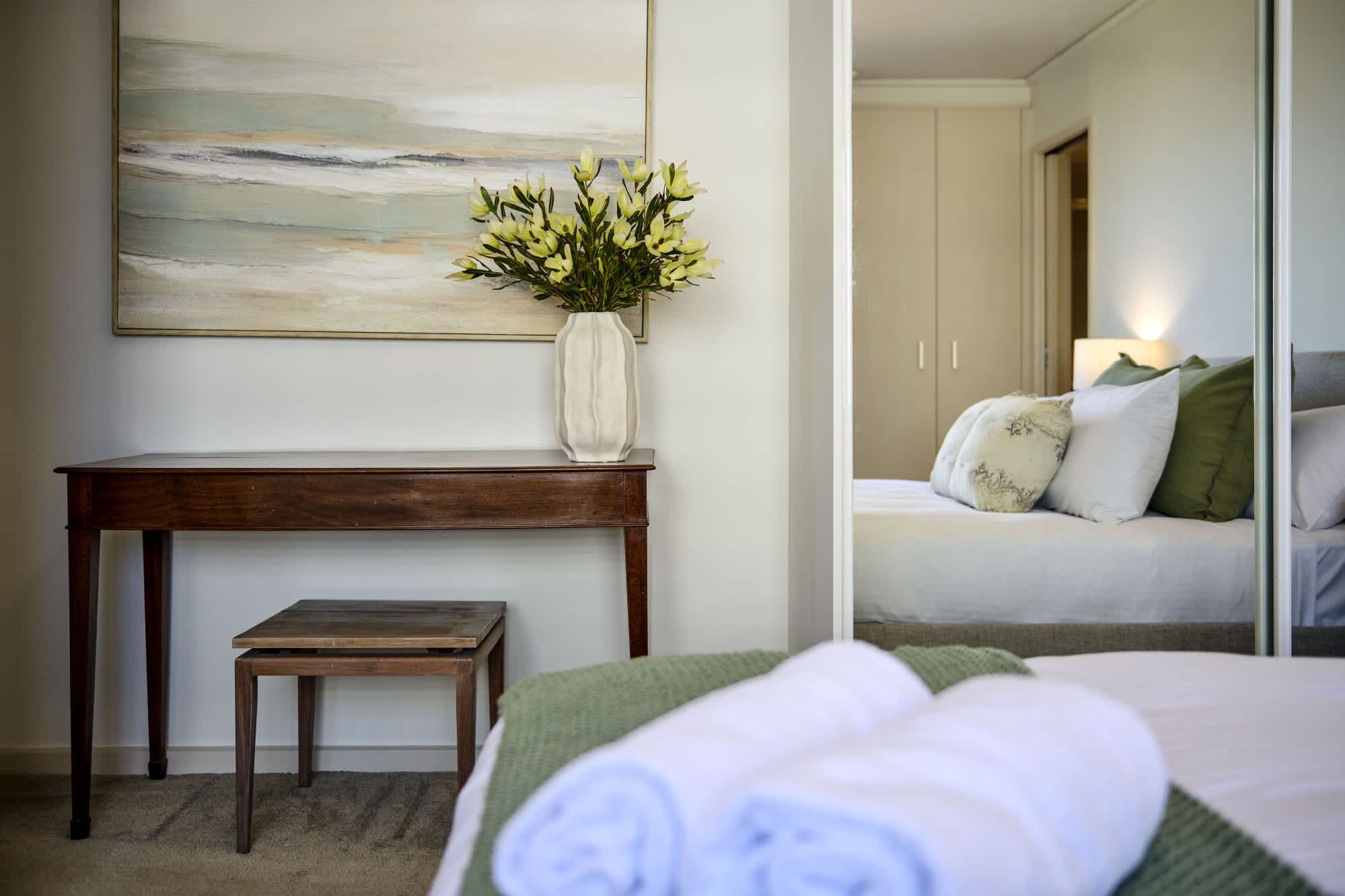 A hotel room with a bed, a small table, a chair, a vase with flowers, and a painting on the wall.