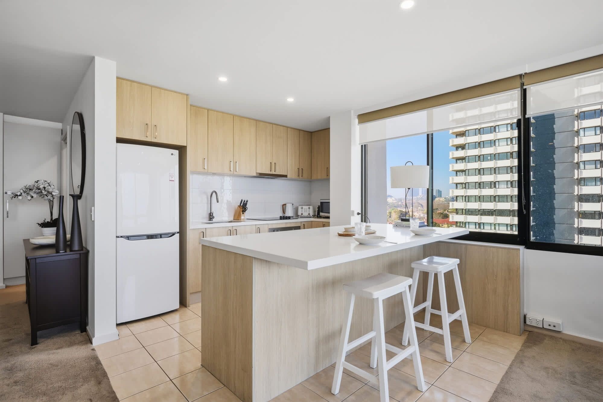 A modern kitchen with a central island, white bar stools, and a view of a cityscape through large windows.