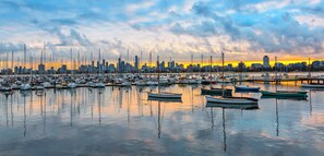 A marina with numerous boats docked, a city skyline in the background, and a sunset. - St Kilda Ocean View with Parking (Saint Kilda West)
