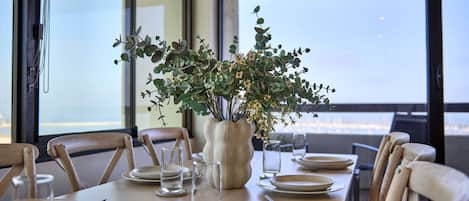 A dining table set for four with a vase of greenery in the center, large windows, and a view of the outdoors.