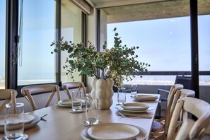 A dining table set for four with a vase of greenery in the center, large windows, and a view of the outdoors. - St Kilda Ocean View with Parking (Saint Kilda West)