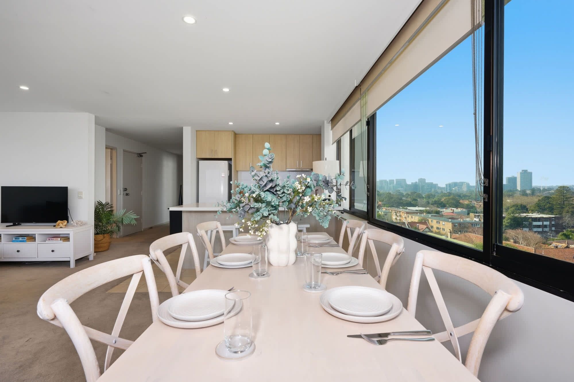 Un espace de repas moderne avec une longue table dressée pour six personnes, un téléviseur dans un coin et une vue sur la ville à travers de grandes fenêtres.