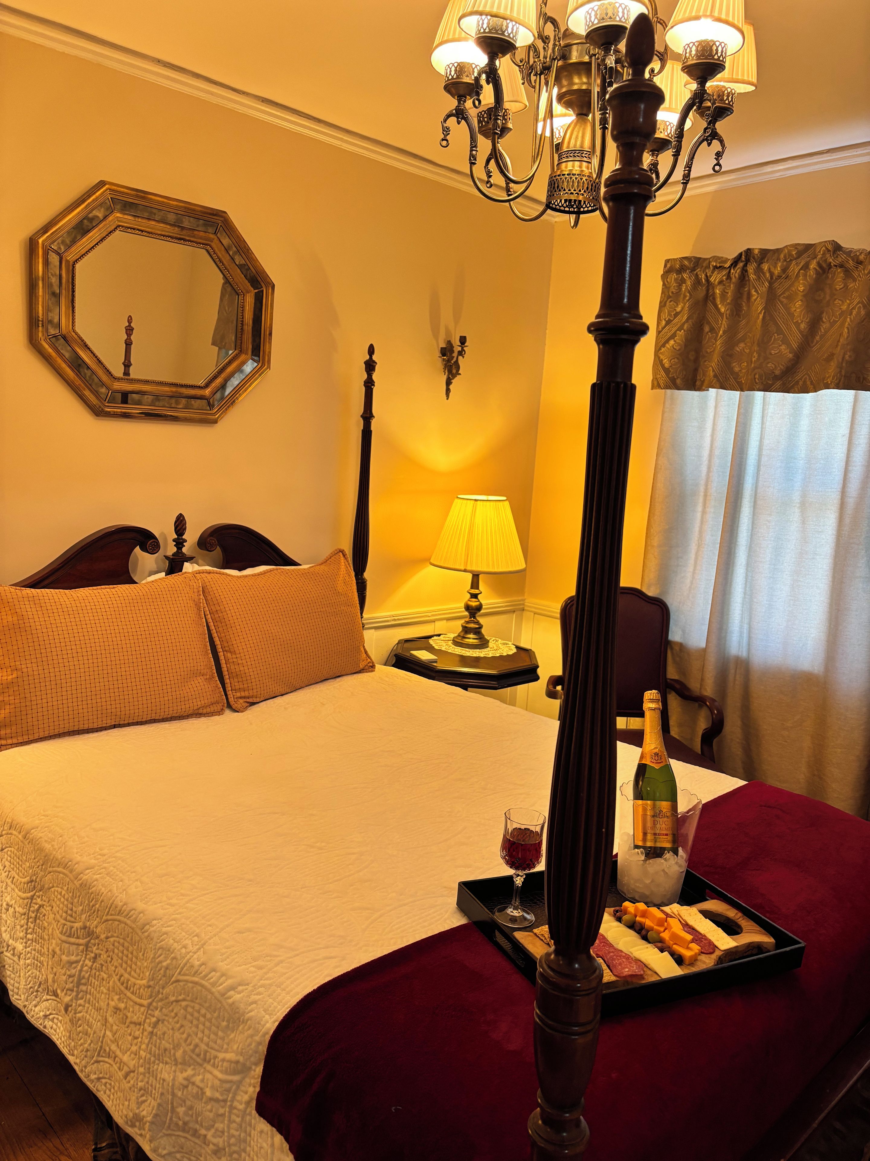 Una habitación de hotel con una cama grande, un candelabro, una mesita de noche con lámpara, una botella de vino y una bandeja de aperitivos.