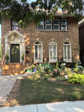 A two-story brick building with a front entrance, a small porch, and a garden area with various plants. - Elegant 1925 Vintage Home (Detroit)