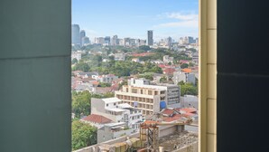 Paisagem urbana com prédios residenciais e comerciais, céu claro e vista pela janela mostrando parte do interior de um edifício.