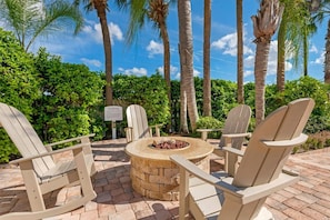 A patio area with Adirondack chairs and a stone fire pit surrounded by lush greenery and palm trees.