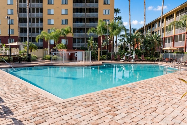 A swimming pool with a brick deck, surrounded by palm trees and a multi-story building with balconies.