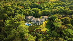 A large, multi-story house surrounded by dense forest, with a swimming pool and a well-maintained lawn.