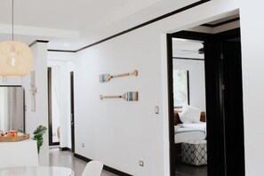 Un interior moderno con zona de comedor, una cama y un banquillo estampado, además de un pasillo con puerta que da a otra habitación.