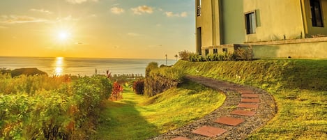 A two-story building with a flat roof and a balcony, situated on a grassy area with a pathway leading to the sea.