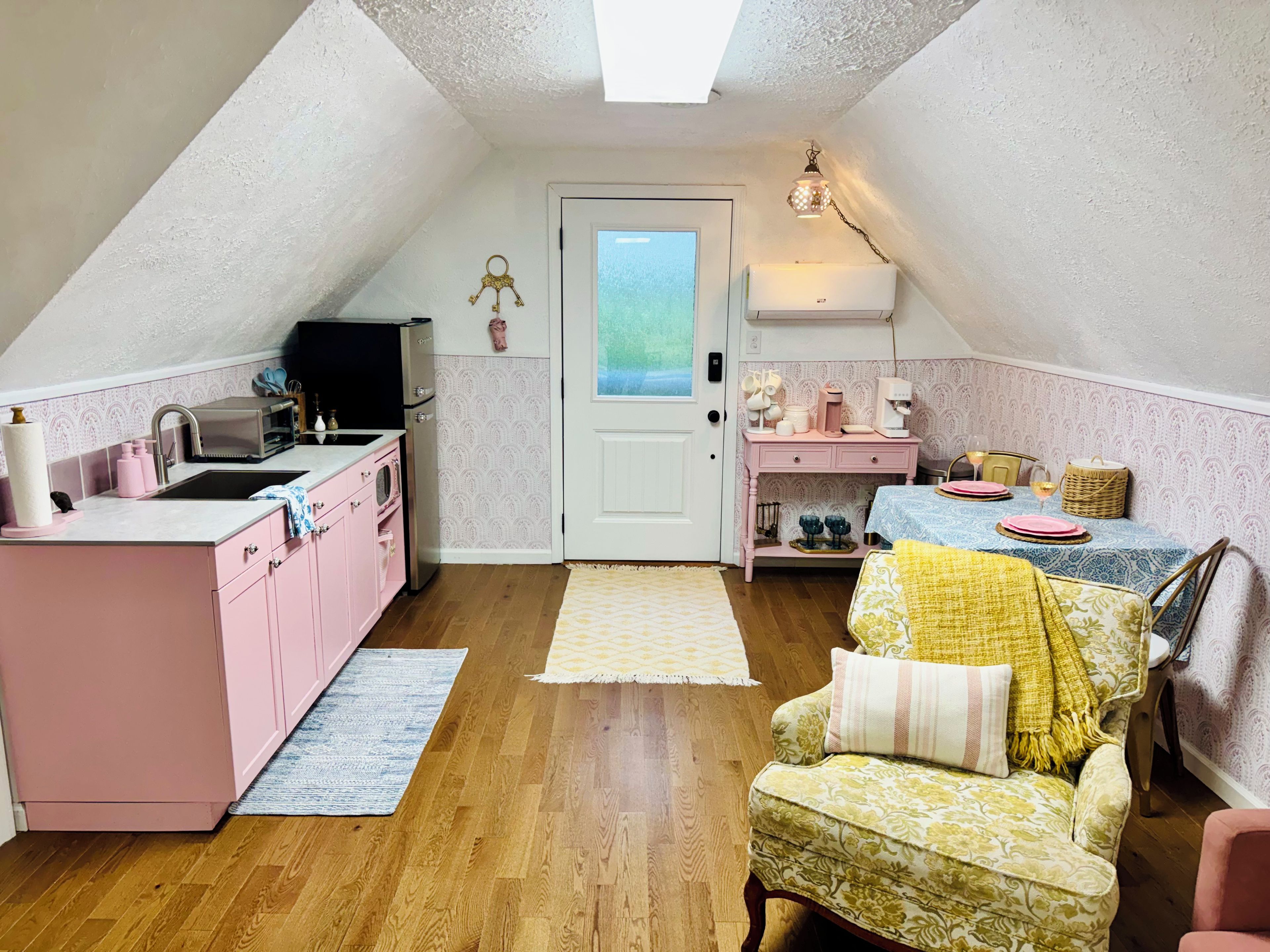 A cozy attic bedroom with a bed, a chair, a desk, and a kitchenette.
