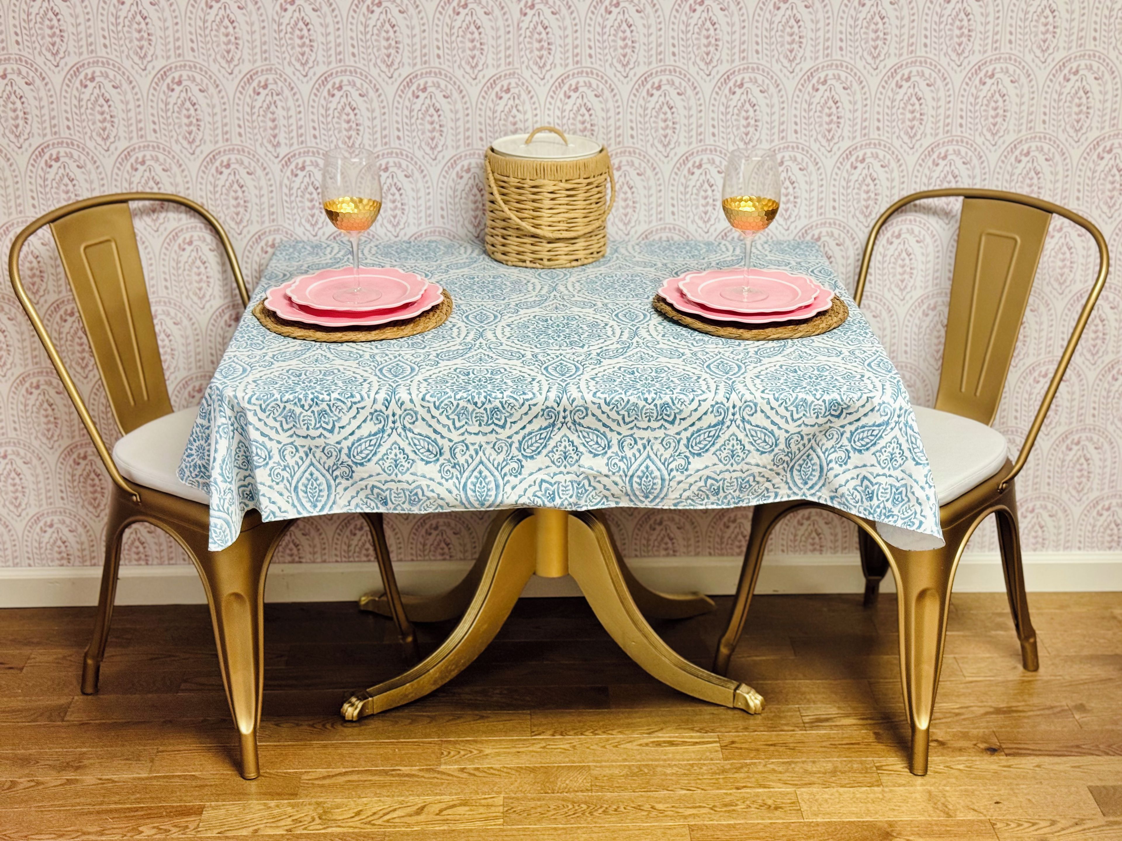 A dining table with a patterned tablecloth, two chairs with gold legs, and a wicker basket on the table.