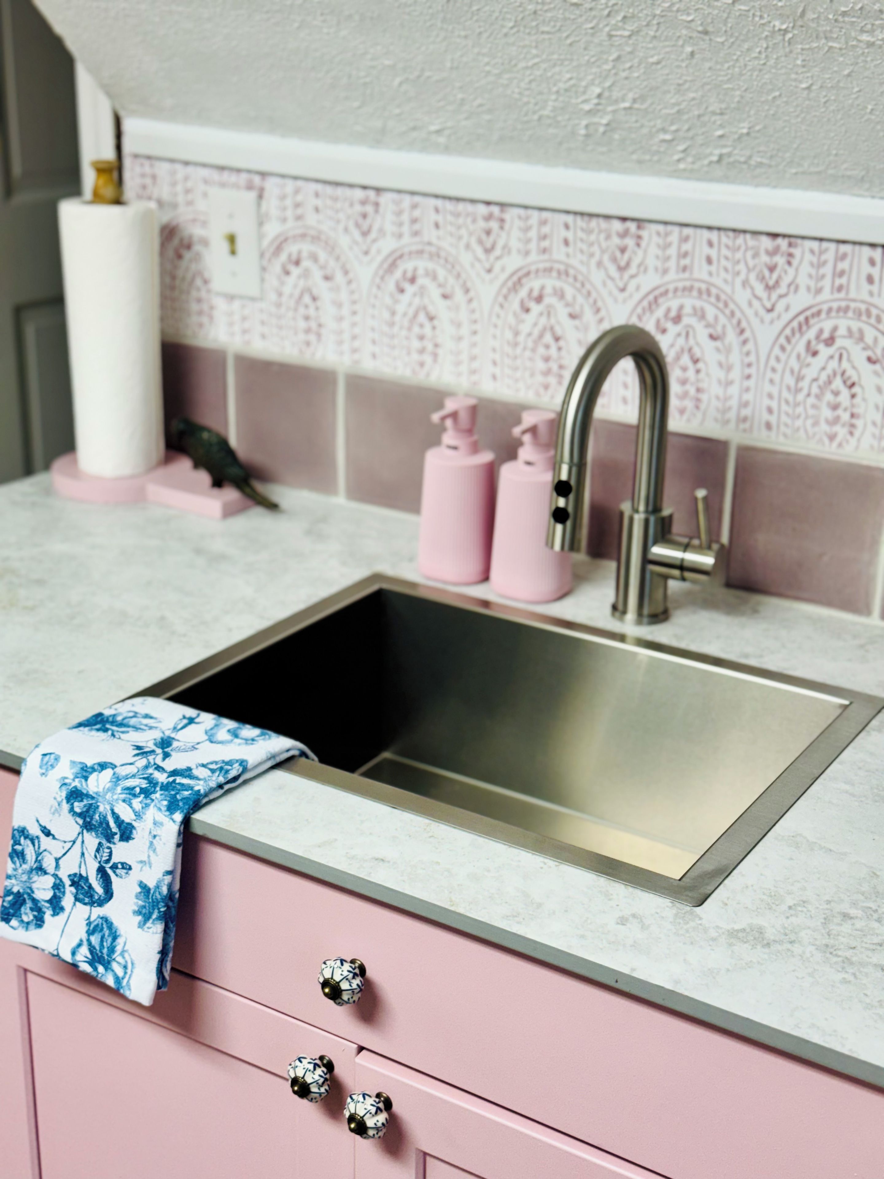 Una cocina con un lavamanos de acero inoxidable, armarios rosados y un salpicadero decorado.