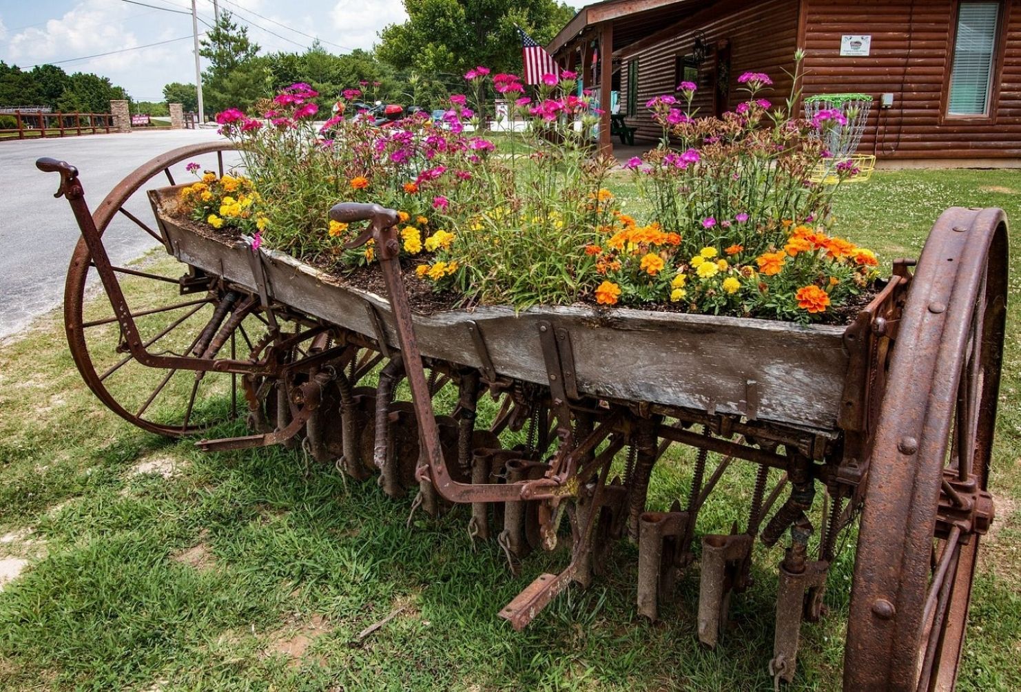 Ein altmodischer Schubkarren, gefüllt mit bunten Blumen, steht auf dem Rasen vor einer Holzhütte.