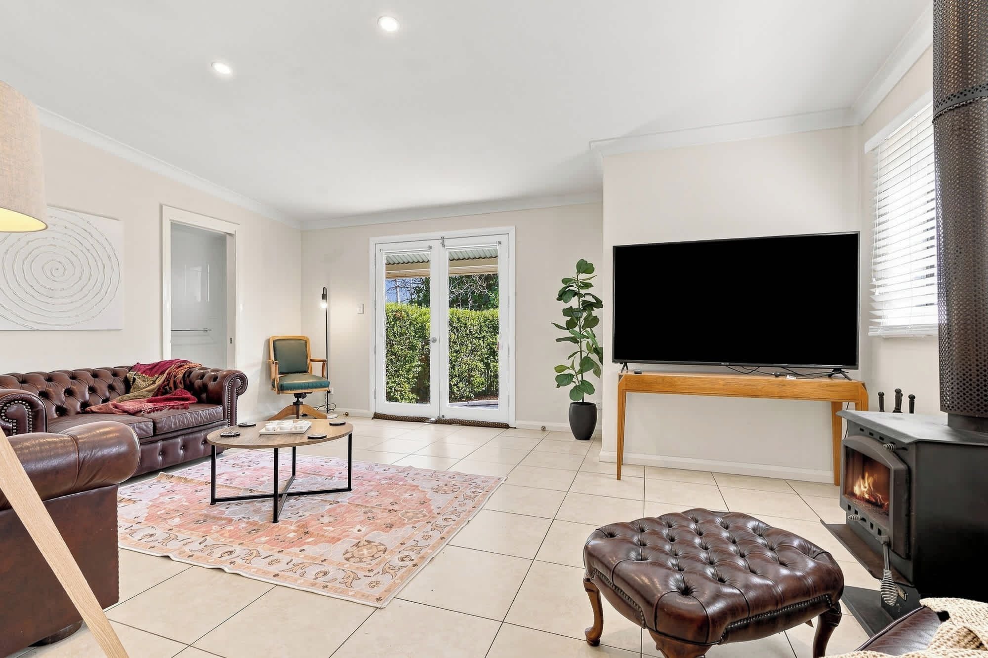 A living room with a leather sofa, a coffee table, a TV on a stand, a fireplace, and a large window with a view of greenery.