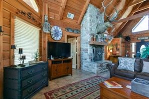 A log cabin interior with a stone fireplace, wooden walls, a television, and a leather sofa. - Branson | Family Nature Getaway | Pool + Disc Golf | 2BR (Ridgedale)