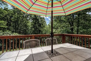 Um pátio com mesa e cadeiras de azulejos, um guarda-sol listrado e um corrimão com vista para uma área arborizada.