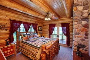A log cabin bedroom with a large bed, wooden headboard, and a stone wall. - Branson | Group Ozarks Retreat | Pools + Fitness | 3BR • Sleeps 8 (Ridgedale)