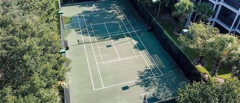 Vue aérienne d’un court de tennis entouré d’une végétation dense et d’un bâtiment doté d’un toit en pente.