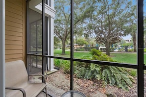 A porch with a chair, a glass door, and a view of a park with trees and a lawn. - Spacious Ground Floor Condo With Pool View! (Davenport)