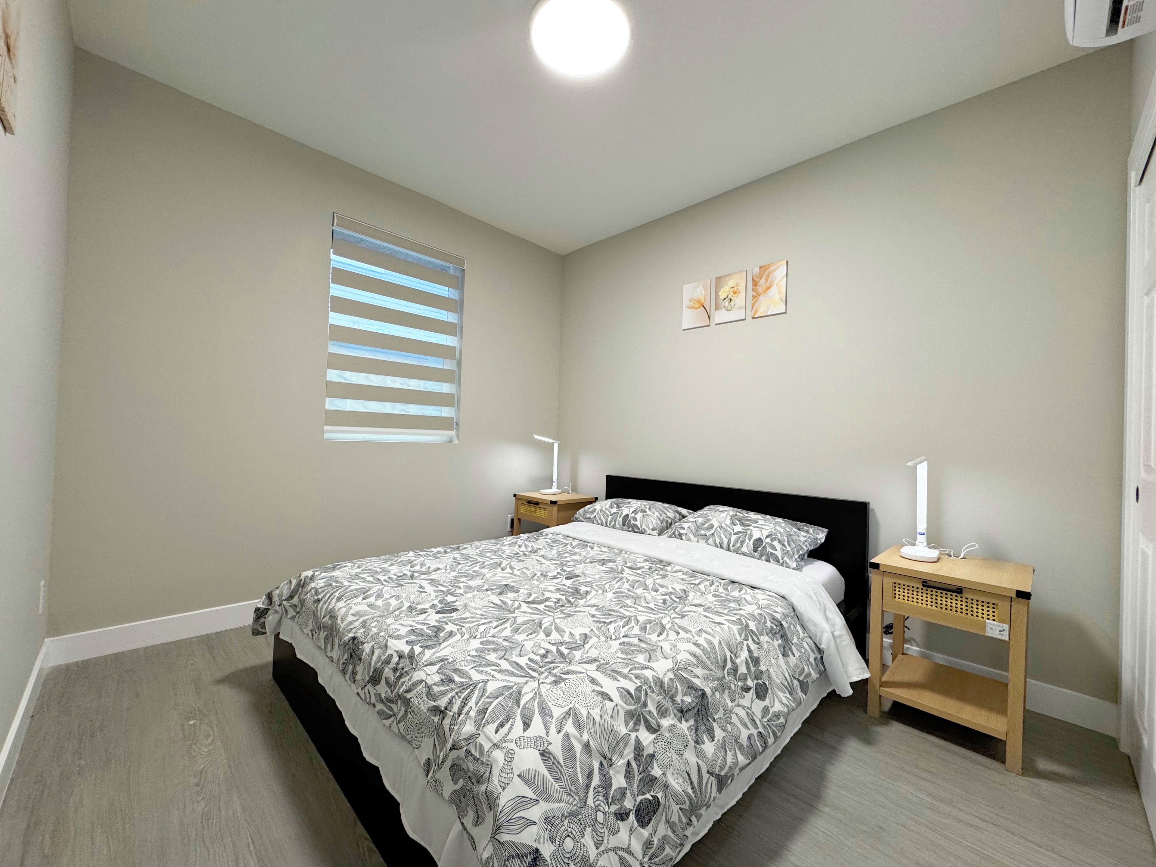 Ein Schlafzimmer mit einem Bett, Nachttischen, einem Fenster mit Jalousien und Wandbildern.