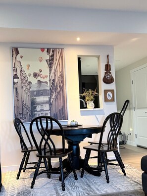 Un coin repas avec une table ronde, quatre chaises, un grand tableau, un miroir, une guitare et un vase rempli de fleurs.