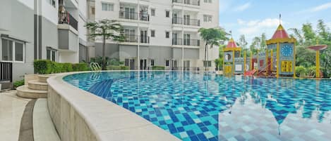 A swimming pool with a checkered pattern, surrounded by a building with balconies and a playground area with a slide and climbing structures.