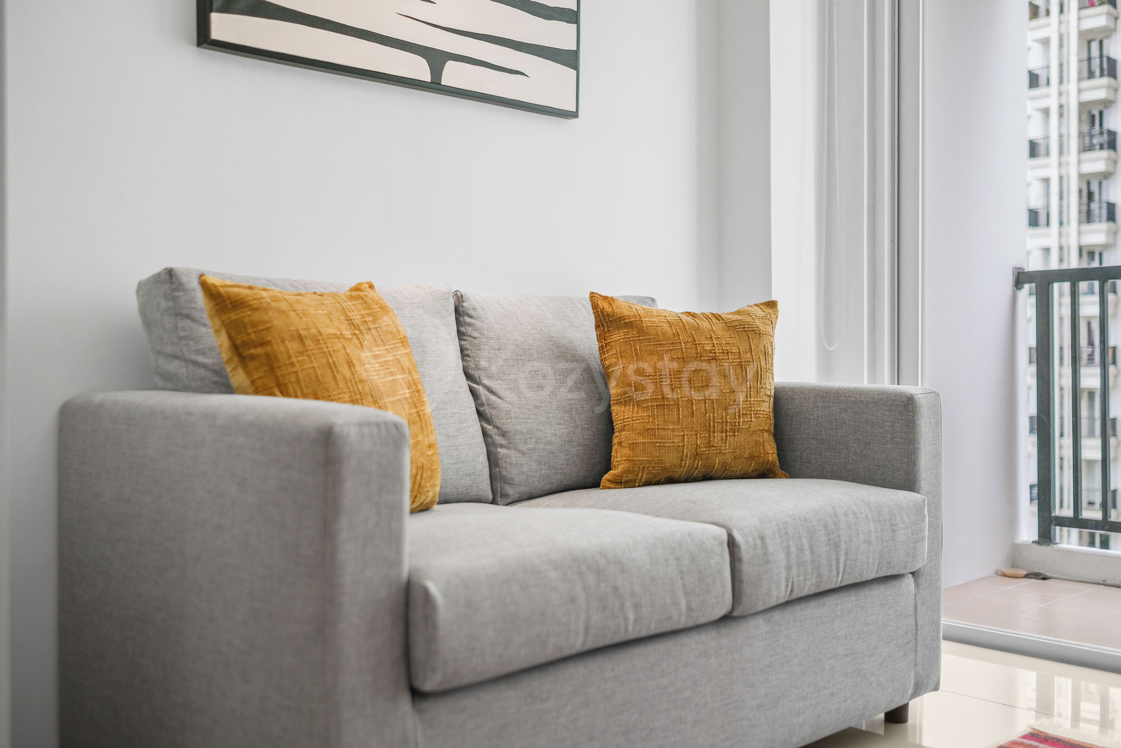 A grey sofa with two mustard-colored cushions against a white wall, with a window and a balcony in the background.
