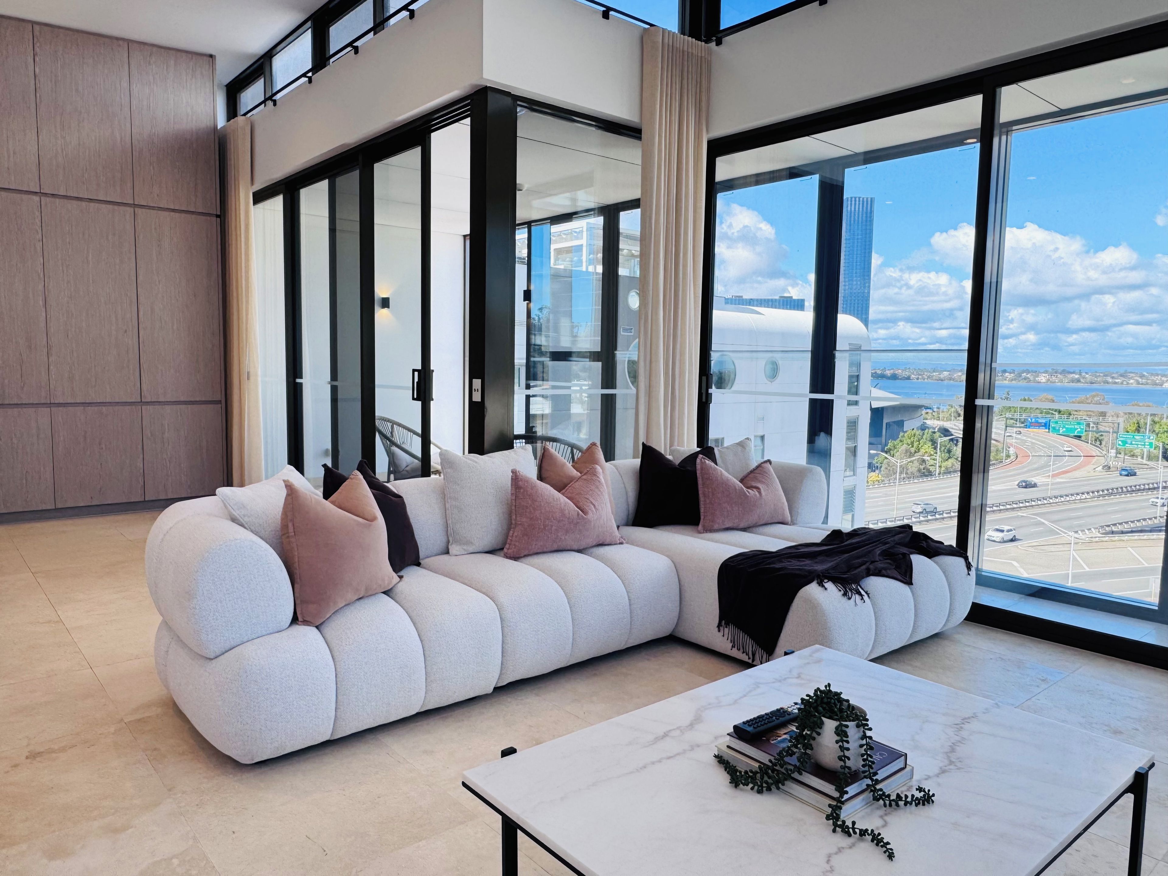 A modern living room with a sectional sofa, a coffee table with books and a plant, large windows with a city view, and a high ceiling with a railing.